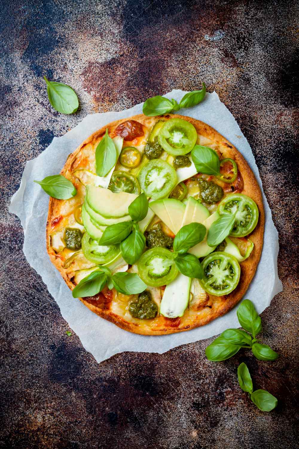 kleine runde Pizza belegt mit grünen Tomaten, Avocadoscheiben und Basilikum